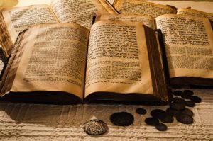Ancient, yellowed books lie open on top of a linen table cloth. They show old Hebrew writing and there are ancient coins on the table.