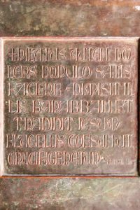A square metal copper-coloured plate is engraved with a verse from Mark 15:15 in Vulgate Latin. The Vulgate is still used today