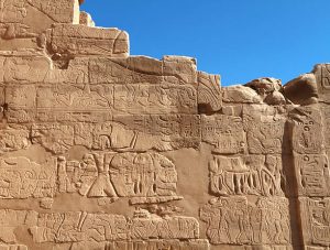 A warm coloured stone wall bearing inscriptions of the peace treaty and drawings showing the history of the Battle of Kadesh against a background of bright blue sky. 